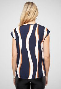 Blouse à manches courtes avec un fond marine et des rayures ondulées orange et blanches en bold. Tissu lisse avec une coupe décontractée et un ourlet arrondi.