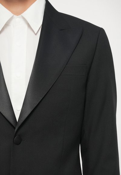 Čierny smokingový sako so saténovými klopami, s jedným čiernym gombíkom a vreckovým štvorcom; spojené s bielou košeľou s okrúhlym golierom.