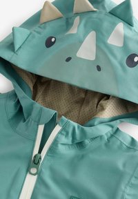 Veste à capuche bleu sarcelle pour enfant avec visage de dinosaure et pics en tissu sur la capuche, doublure intérieure beige et fermeture éclair à l'avant.