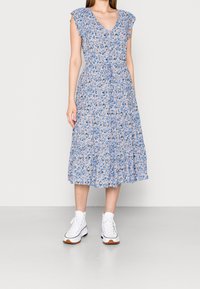 Florales Midi-Kleid mit blauem Hintergrund, featuring Rüschensäulen, einem V-Ausschnitt, elastischem Taillenbund und gestuftem Rock. Getragen mit weißen Sneakers.