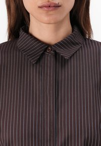 Chemise rayée marron avec des fines rayures bleu clair, un col pointu et un unique bouton marron en haut. Texture de tissu lisse.