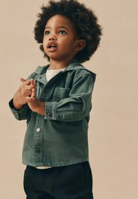 Veste en denim verte avec capuche, dotée d'une fermeture à boutons-pression et d'une poche poitrine, portée par-dessus une chemise blanche et associée à un pantalon noir.