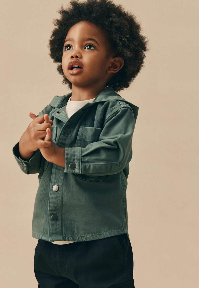 Veste en denim verte avec capuche, dotée d'une fermeture à boutons-pression et d'une poche poitrine, portée par-dessus une chemise blanche et associée à un pantalon noir.