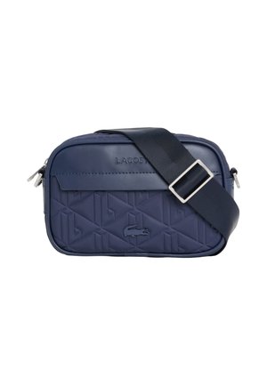 Bolso bandolera rectangular azul marino con correa negra ajustable, patrón geométrico en relieve, logo de Lacoste y compartimentos con cremallera.