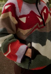 Pullover in pile con zip a quarto, caratterizzato da un motivo geometrico in rosso, verde, panna e viola, con una finitura testurizzata e soffice.
