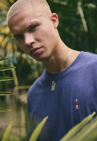 Blue cotton t-shirt featuring a small embroidered ketchup bottle on the left chest, worn with a silver necklace and surrounded by green foliage.