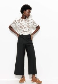 Haut en dentelle florale blanche associé à un pantalon large noir et des baskets en cuir texturé marron. Les accessoires incluent des boucles d'oreilles et des bracelets en or.