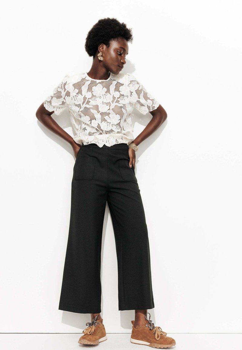 Haut en dentelle florale blanche associé à un pantalon large noir et des baskets en cuir texturé marron. Les accessoires incluent des boucles d'oreilles et des bracelets en or.