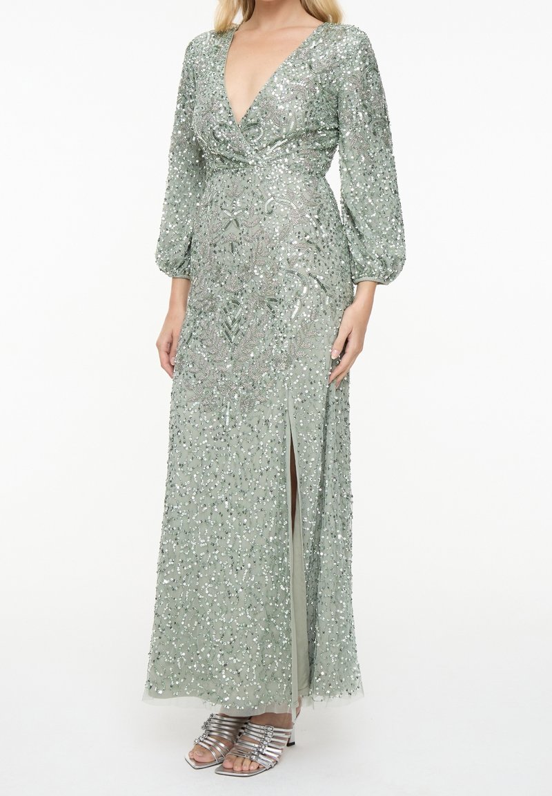 Longue robe vert clair ornée de sequins, manches longues bouffantes, décolleté en V profond, fente latérale, portée avec des talons argentés à brides.