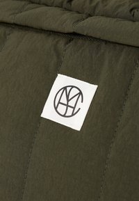 Olivgrüner gesteppter Stoff mit einer strukturierten Oberfläche, versehen mit einem weißen Etikett mit einem schwarzen Logo. Nahtdetails sind sichtbar.