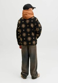 Crna flis jakna s uzorkom i smešnim kapuljačom, uparena s labavim sivim hlačama i bež obućom. Nošena crna kapa ravnog dna.