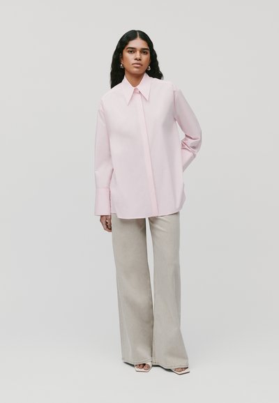 Camicia oversized rosa chiaro con colletto a punta, abbinata a pantaloni a palazzo beige chiaro. Tessuto morbido e vestibilità rilassata.