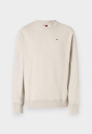 Beżowa longsleeve bluza crewneck z prążkowanymi mankietami i dolną częścią, z małym czerwono-biało-niebieskim logo flagi na piersi.