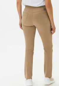 Pantalons stretch beiges avec une texture lisse, dotés de deux poches arrière et d'une taille élastique. Associés à des chaussures blanches.