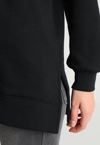 Mano appoggiata accanto all'anca, indossando una maglietta nera a maniche lunghe con fessura laterale sopra pantaloni grigi su uno sfondo neutro.