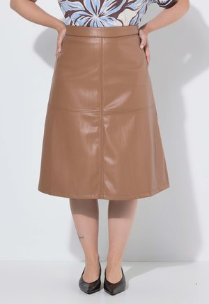 Femme portant une jupe en cuir marron longueur genou, un haut fleuri, des talons pointus noirs, debout, mains sur les hanches, sur fond blanc.