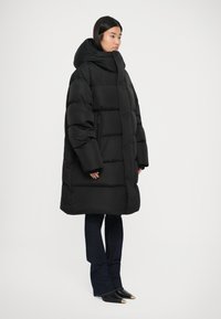 Abrigo de plumas negro con capucha extragrande, diseño acolchado, bolsillos laterales con cremallera y mangas anchas; llevado sobre unos vaqueros acampanados azules y botas de punta negra.