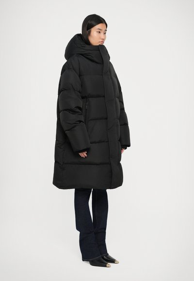 Cappotto puffer nero con cappuccio oversize, design trapuntato, tasche laterali con zip e maniche ampie; indossato sopra jeans a zampa d'elefante blu e stivali neri a punta.