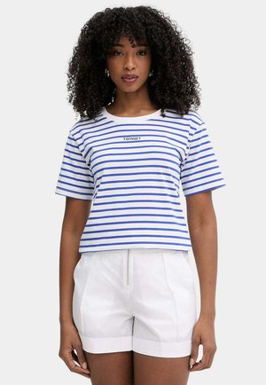 Giovane donna con capelli ricci che indossa una T-shirt bianca e blu a righe con l’etichetta "TWINSET" e shorts bianchi a vita alta.