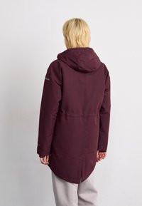 Person mit kurzen blonden Haaren, die eine weinroten Winterjacke mit Kapuze und hellfarbene Hose trägt und mit dem Rücken zu einem schlichten Hintergrund steht.