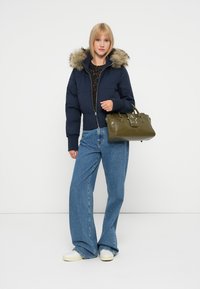 Geacă puffer bleumarin cu glugă din blană sintetică, purtată cu un top negru cu model și blugi largi albaștri. Geantă de mână kaki cu detalii în formă de stea.