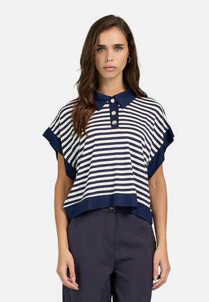 Donna che indossa una polo a righe blu navy e bianco a maniche corte con tre bottoni, abbinata a pantaloni blu navy scuro, in piedi davanti a uno sfondo semplice.