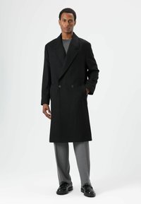 Zwarte dubbelgeknoopte jas met brede revers, zijzakken, gedragen over een grijs shirt, gecombineerd met grijze broek en zwarte schoenen.