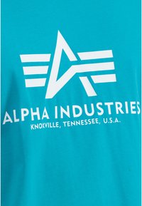 Tyrkysové bavlnené tričko s bielym textom: "ALPHA INDUSTRIES" a "KNOXVILLE, TENNESSEE, U.S.A." spolu so stylizovanou hviezdovou logom.