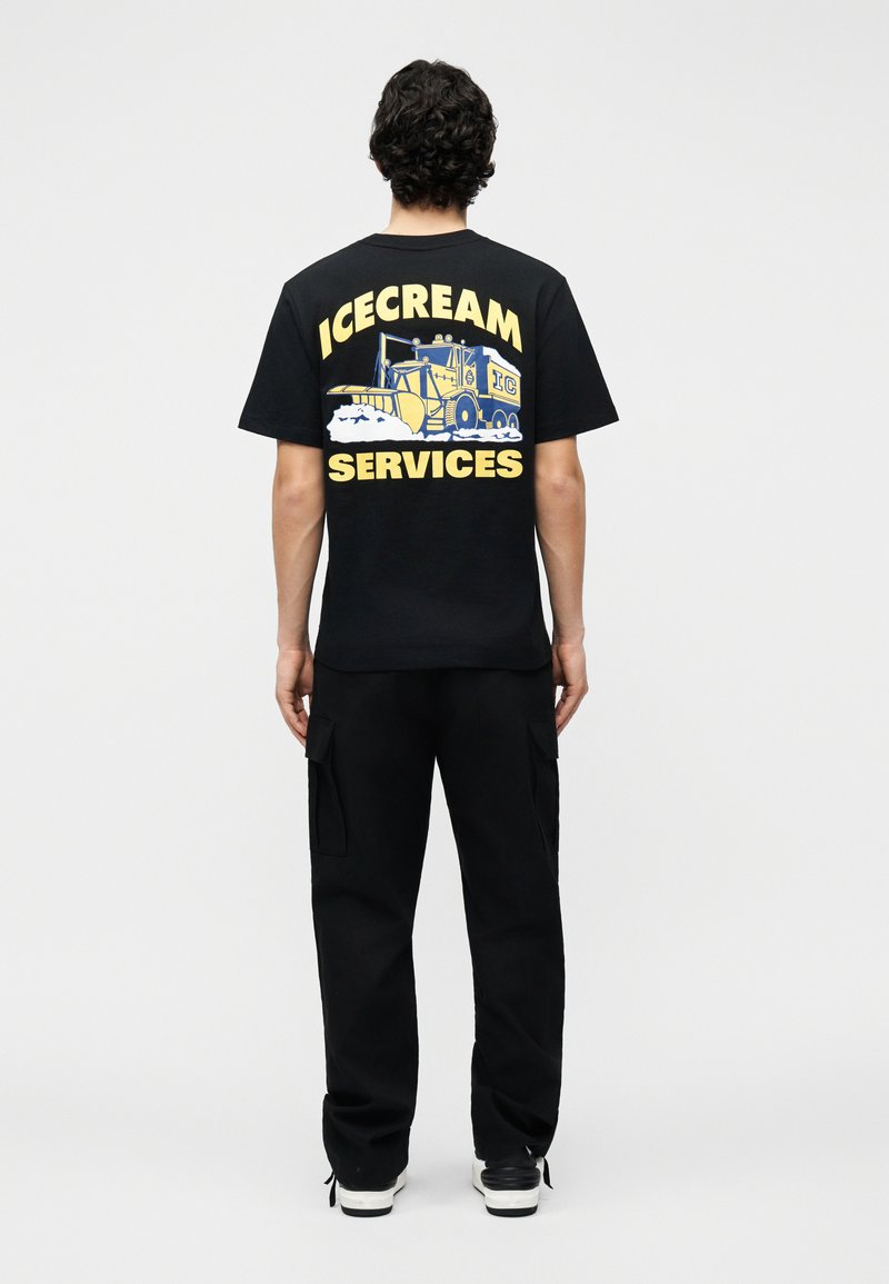 Άνδρας φοράει μαύρο T-shirt με κίτρινο και μπλε παγωτατζίδικο και τις λέξεις «ICECREAM SERVICES» στην πλάτη, μαύρο παντελόνι και μαύρα-λευκά παπούτσια.