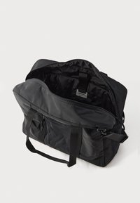 Crna mekana platnena torba za duffel s dvostrukim ručkama, prednjim zip džepom, podešivim remenom za rame i otvorenom glavnom pregradom koja prikazuje unutrašnju oznaku.