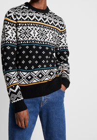 Pull en maille noir avec un motif géométrique blanc, turquoise et doré. Encolure et poignets côtelés, tissu texturé. Porté avec un jean bleu.