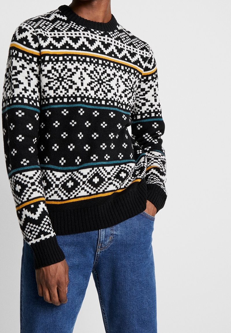 Pull en maille noir avec un motif géométrique blanc, turquoise et doré. Encolure et poignets côtelés, tissu texturé. Porté avec un jean bleu.
