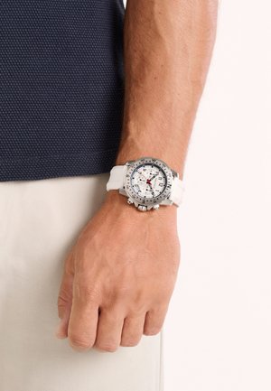 Montre chronographe argentée avec un bracelet en silicone blanc, cadran rond avec des marquages noirs et une trotteuse rouge, portée au poignet.