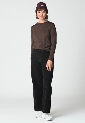 Femme debout portant un bonnet bordeaux, une chemise marron à manches longues, un pantalon noir et des baskets blanches sur un fond clair uni.