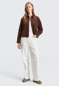 Bruine corduroy knoopshirt, witte t-shirt, wijde witte broek en beige schoenen. Model staat tegen een effen lichte achtergrond.