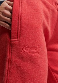 Hand holding the edge of a red SuperDry hoodie pocket showing embroidered logo and Japanese text on soft fabric.