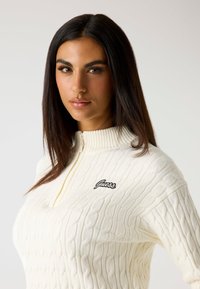 Maglione a trecce color crema con colletto a coste e design con mezza zip; presenta il logo nero "Guess" sul lato sinistro.