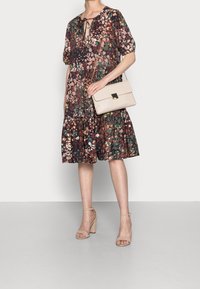 Abito floreale con maniche corte a palloncino, scollatura a chiave e gonna a strati. Modella che tiene una borsa leggera beige. Tacchi beige.