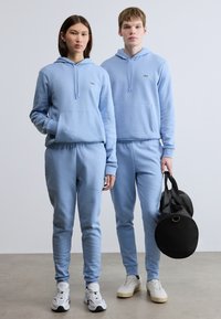 Φωτεινά γαλάζια φούτερ με κουκούλα και αντίστοιχα παντελόνια φόρμας από μαλακό ύφασμα, με τσέπες και μικρό λογότυπο. Το μοντέλο κρατά μια μαύρη τσάντα.