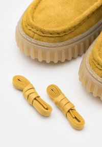 Gelbe Wildleder-Schuhoberteile mit durchsichtigen Gummisohlen, begleitet von zwei Sets passender gelber Schnürsenkel auf einem weißen Hintergrund.