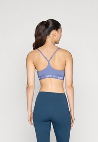 Sujetador deportivo morado con tirantes ajustables, que presenta una amplia banda con el logo de Nike en la parte inferior; combinado con leggings de tiro alto en un azul oscuro.
