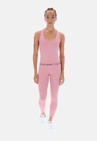 Prilega roza športna majica brez rokavov in legice z črnim logotipom na pasu, v kombinaciji z belimi športnimi čevlji. Gladek, raztegljiv material.