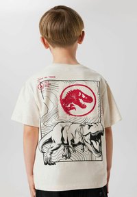 Camiseta color crema con un diseño gráfico de un dinosaurio rugiente, bordeado por un emblema circular rojo y líneas de terreno.