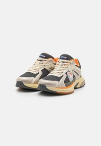 Sneaker beige e nere con pannelli in rete, accenti grigi, dettagli arancioni e un logo bianco. Presentano una suola imbottita e una presa testurizzata.