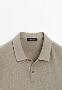 Polo beige à manches courtes avec deux boutons et un col, affichant une étiquette noire avec les textes "Massimo Dutti" et "Made in Cambodia".