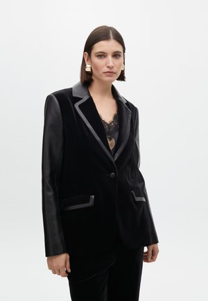 Blazer in velluto nero con dettagli in pelle, rever a punta e tasche frontali. Presenta una chiusura a un bottone e una texture liscia.