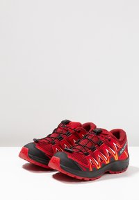 Red and black athletic shoes featuring a mesh upper, elastic lacing system, and a rugged outsole. Prominent logo and dynamic design elements.