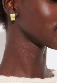 Gold-tone hoop earring features a textured design with circular patterns, showcasing a polished finish against dark skin and a knit sweater.