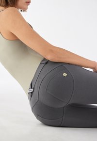 Harmaat urheiluhousut, joissa on sileä pinta, kaarevat taskut ja takana pieni kultainen logo. Vyötärökuminauha on joustava mukavuutta varten.