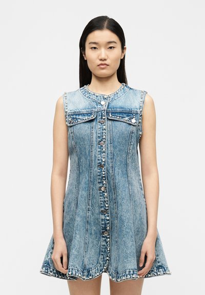 Νεαρή γυναίκα που φορά αμάνικο ανοιχτό μπλε φόρεμα από τζιν με διακοσμητικά πέρλες και κουμπιά μπροστά, σε απλό λευκό φόντο.